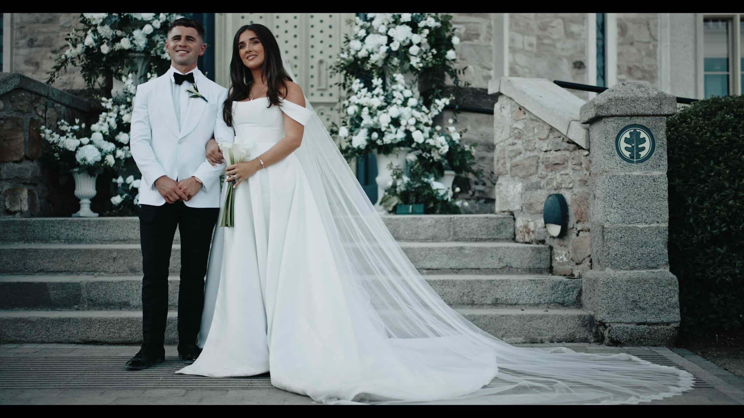 Bride and groom in white attire standing on stone steps with floral decor.