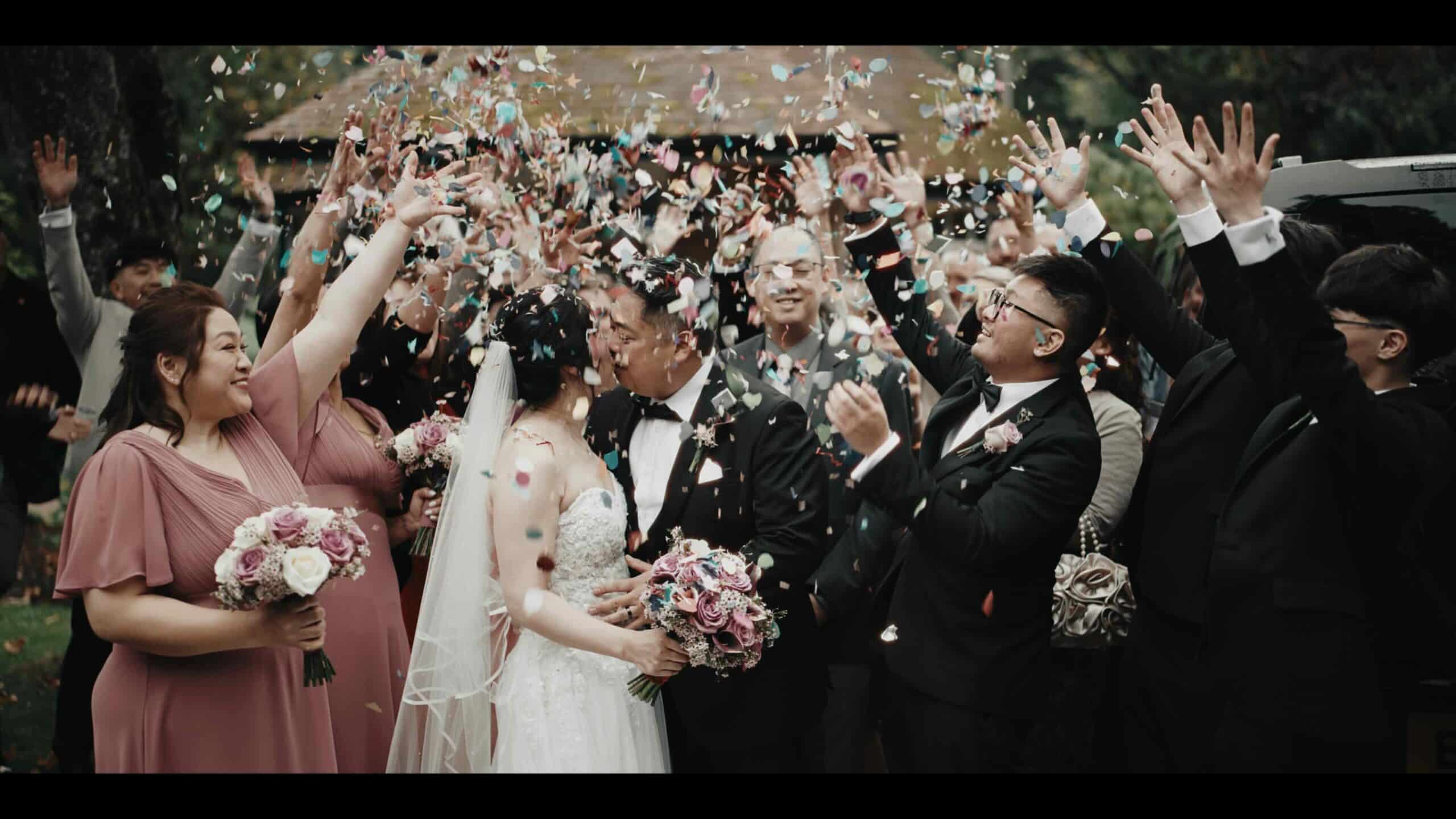 Bride and groom kissing surrounded by joyful guests throwing confetti at a wedding celebration.