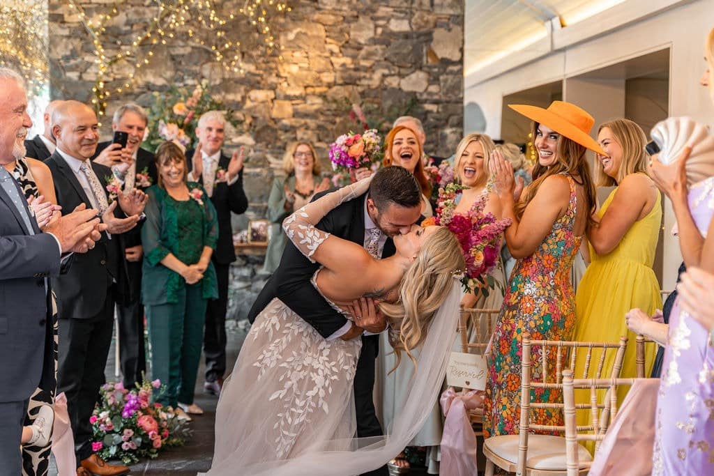 Bride and groom share a romantic kiss at their wedding ceremony, surrounded by applauding guests.