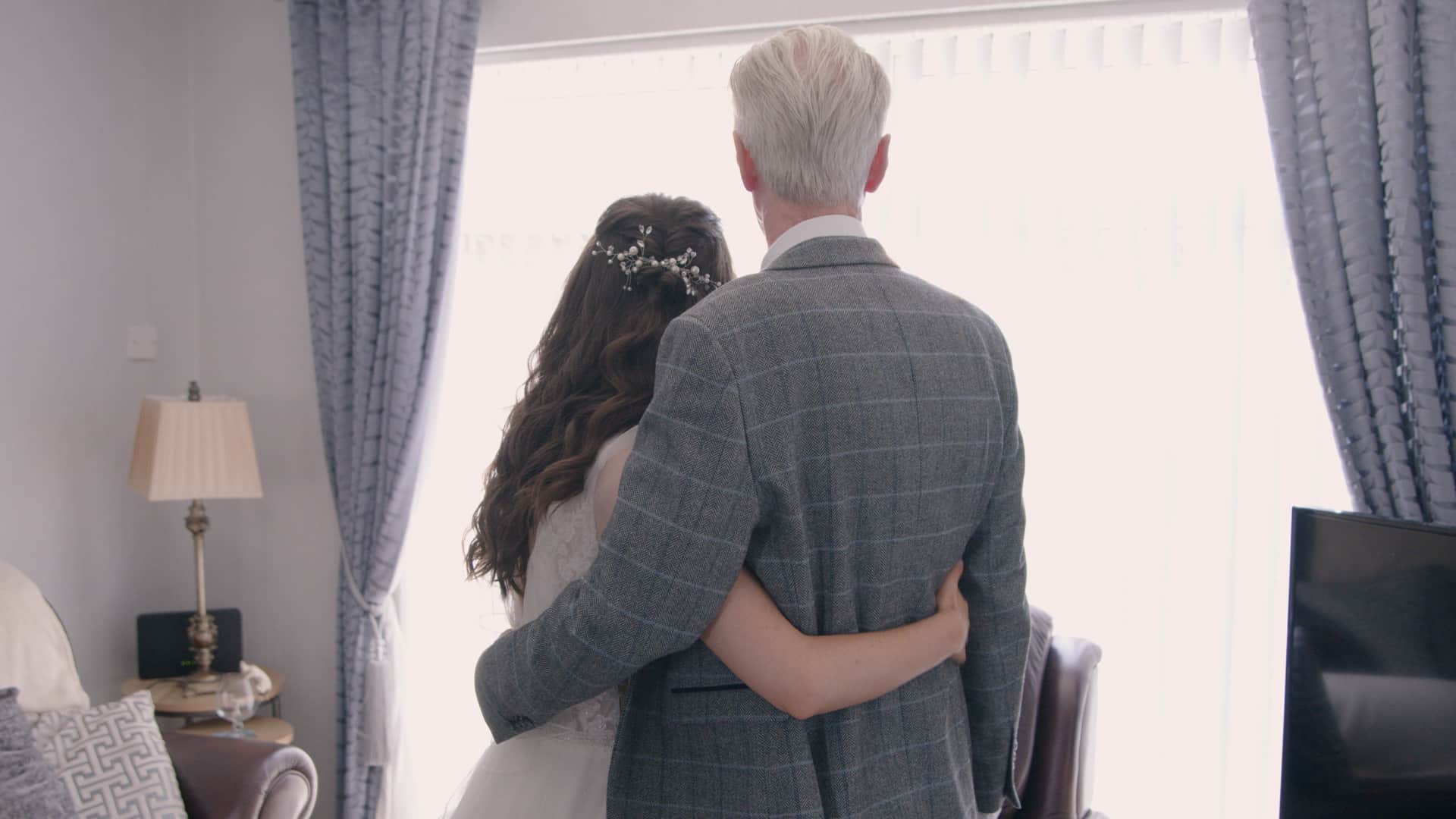 A bride and an older man with white hair stand arm in arm, facing a bright window in a cozy room.