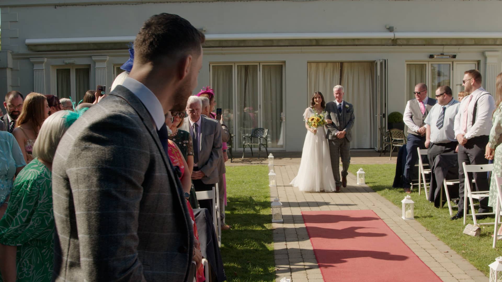 Bride walking down the aisle with her father, surrounded by guests at an outdoor wedding ceremony.