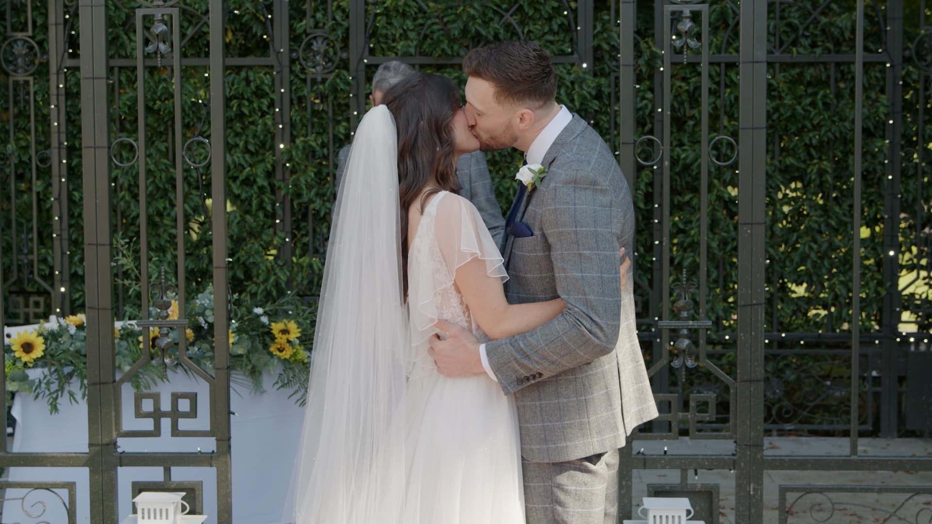 Bride and groom kissing outdoors by a decorative metal gate with sunflowers.