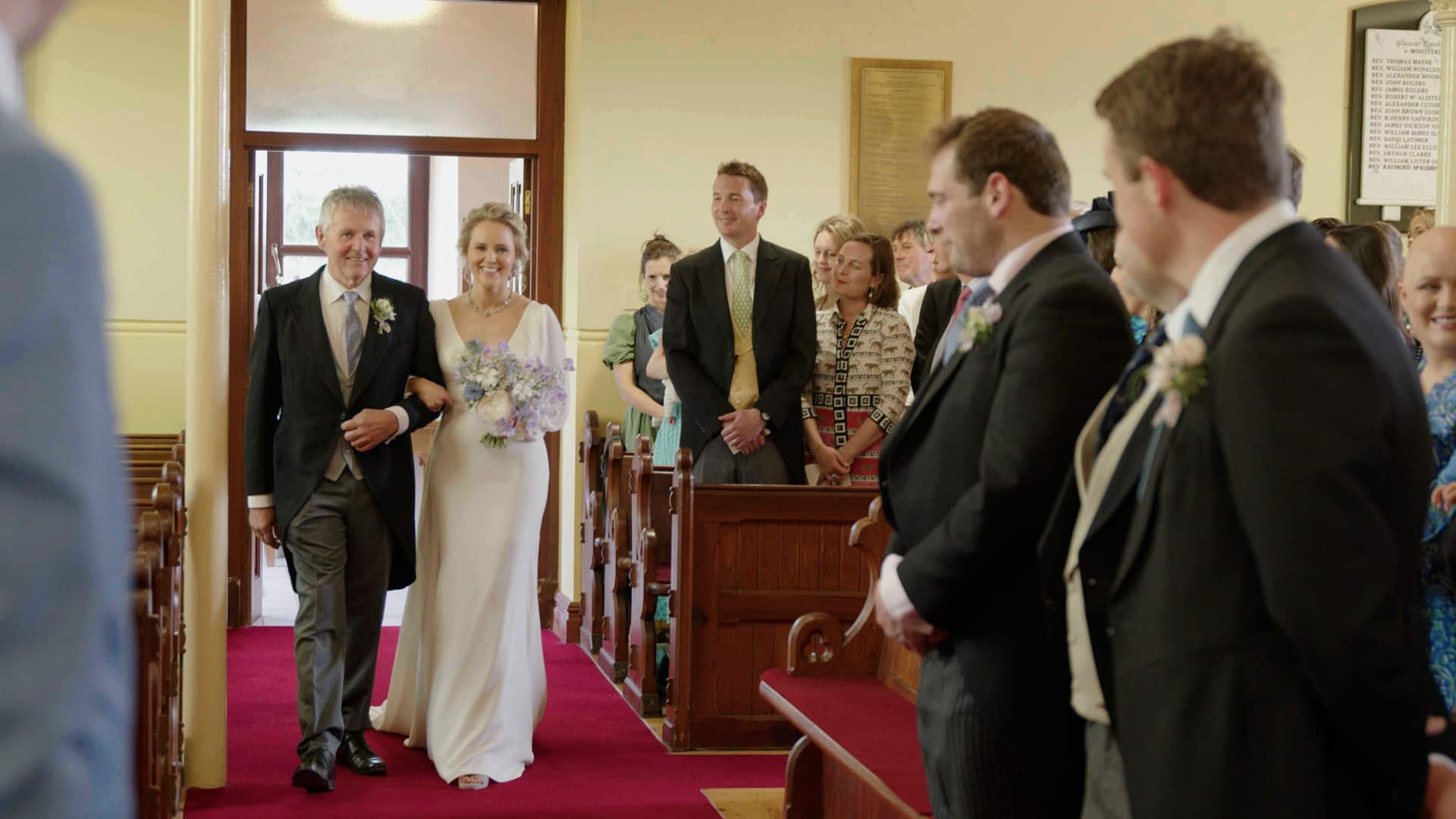 Bride walking down the aisle with her father at a wedding ceremony, guests standing and smiling.
