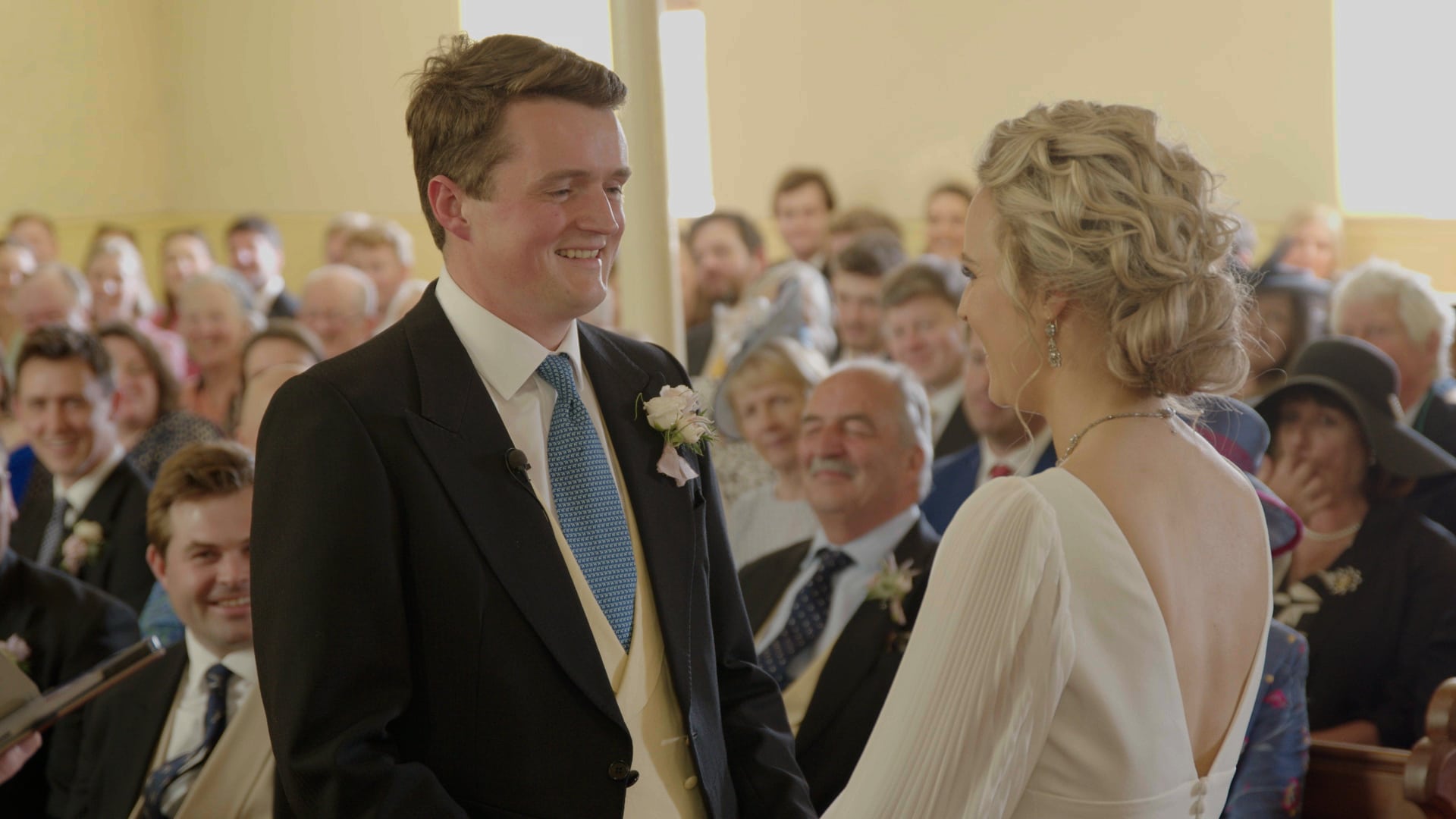 Bride and groom smiling at each other during a wedding ceremony, surrounded by guests.