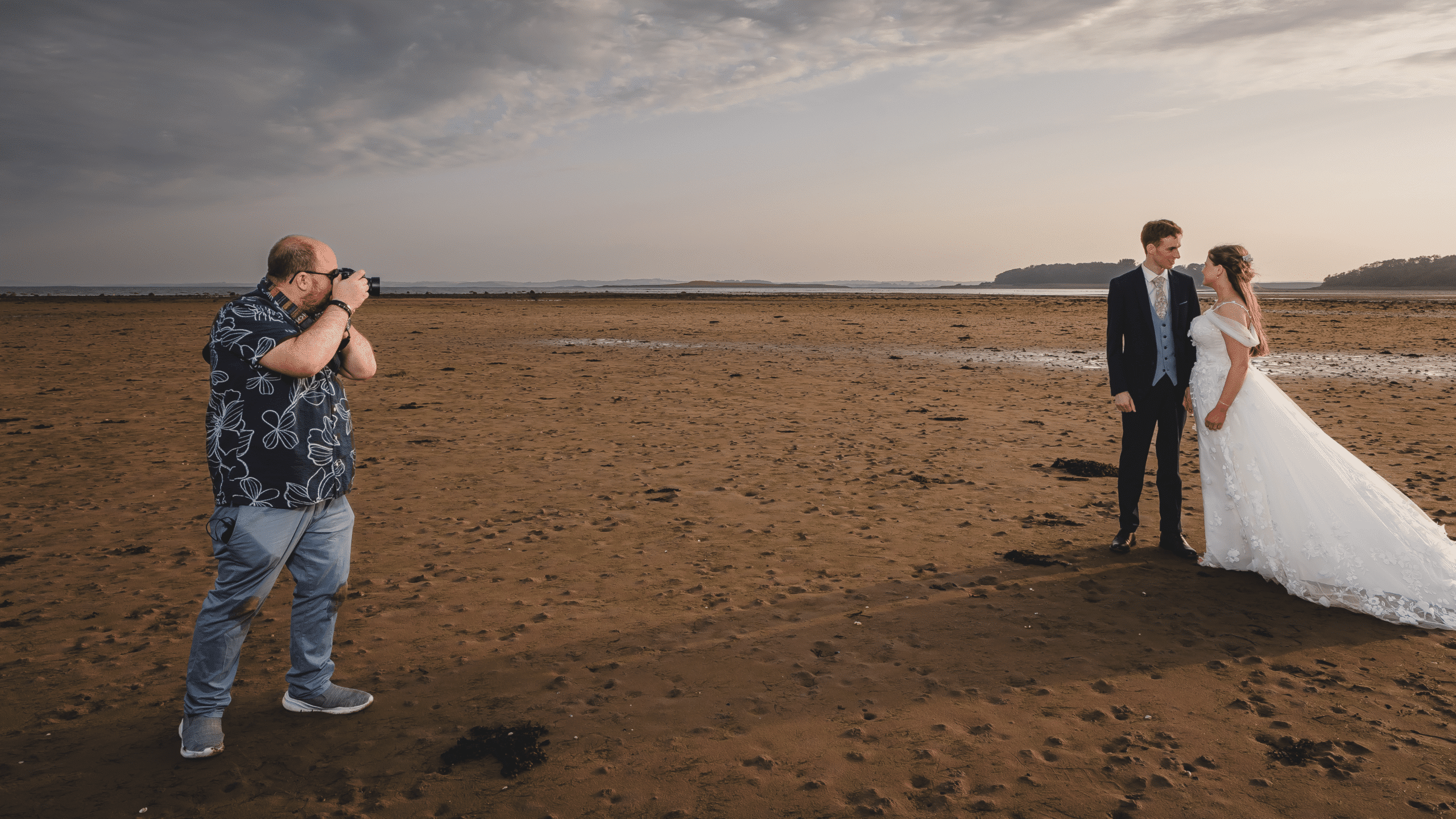 Photographer taking pictures of a couple in wedding attire on a sandy beach at sunset.