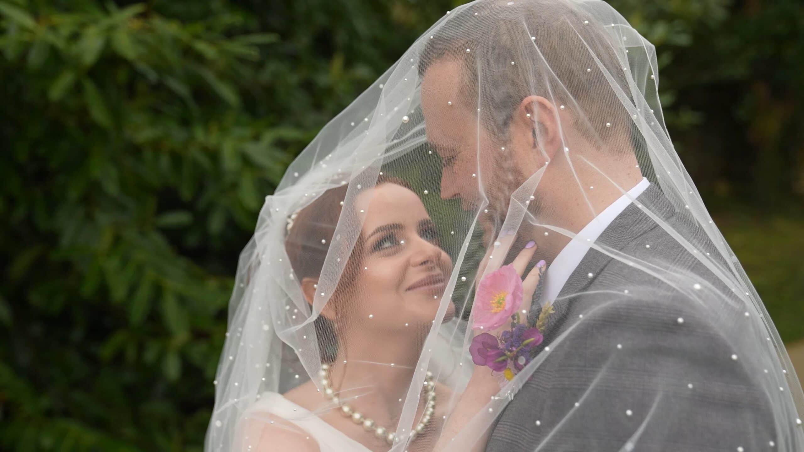 Bride and groom smiling under a sheer, pearl-decorated veil outdoors.