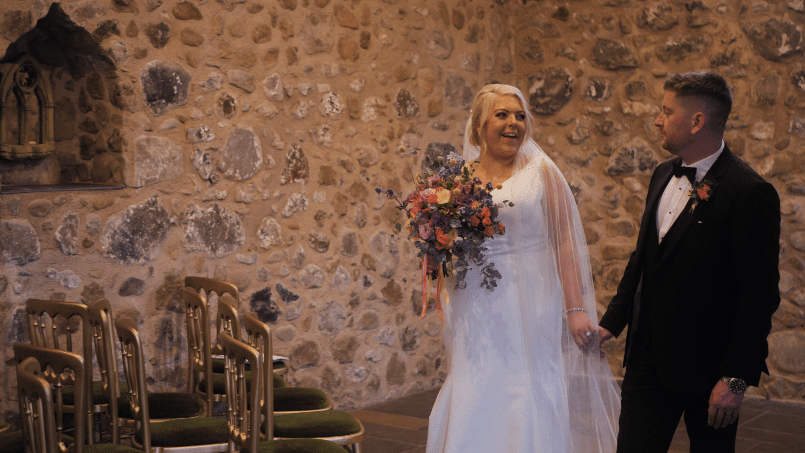 Bride and groom walking in stone-walled venue, bride holding colorful bouquet.