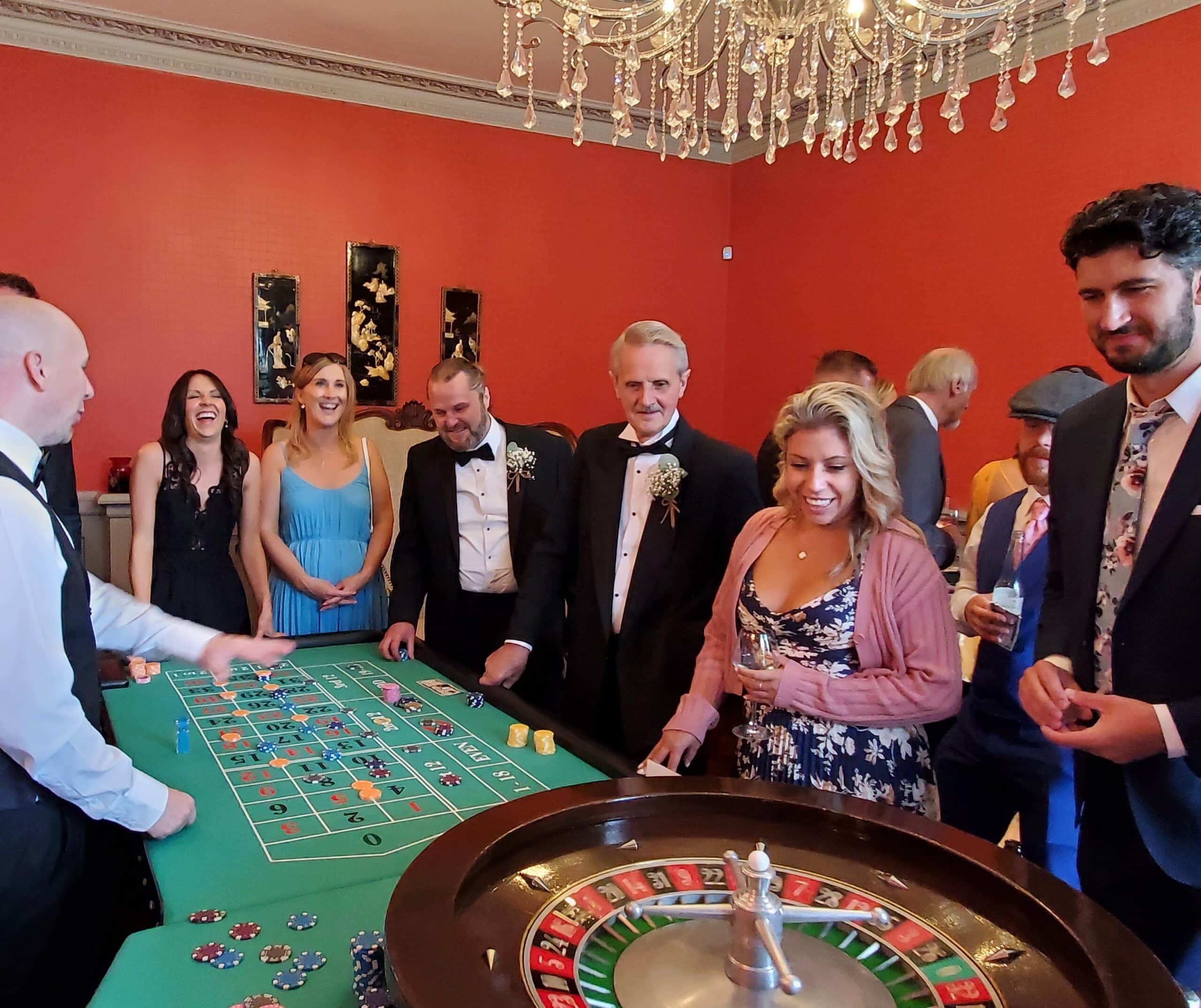 Wedding guests enjoying a roulette table at a reception, set in a red-walled room with a chandelier.