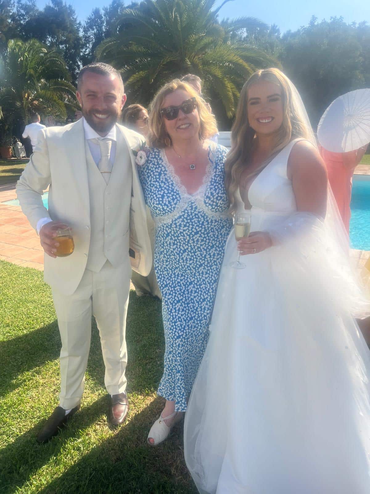 Bride and groom posing with a woman in sunglasses outdoors by a pool on a sunny day.