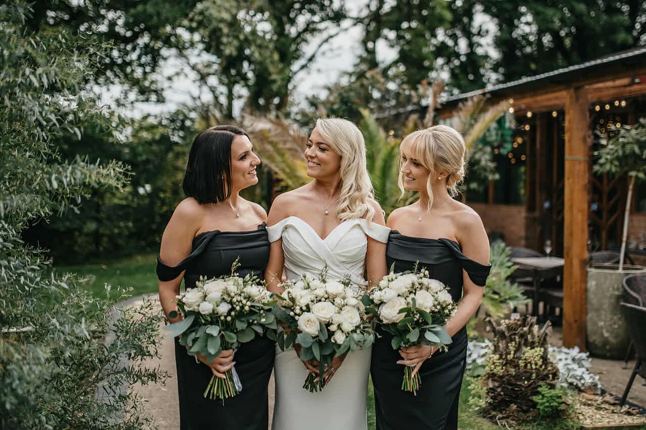 Bride with bridesmaids holding bouquets, standing outdoors, smiling at each other.