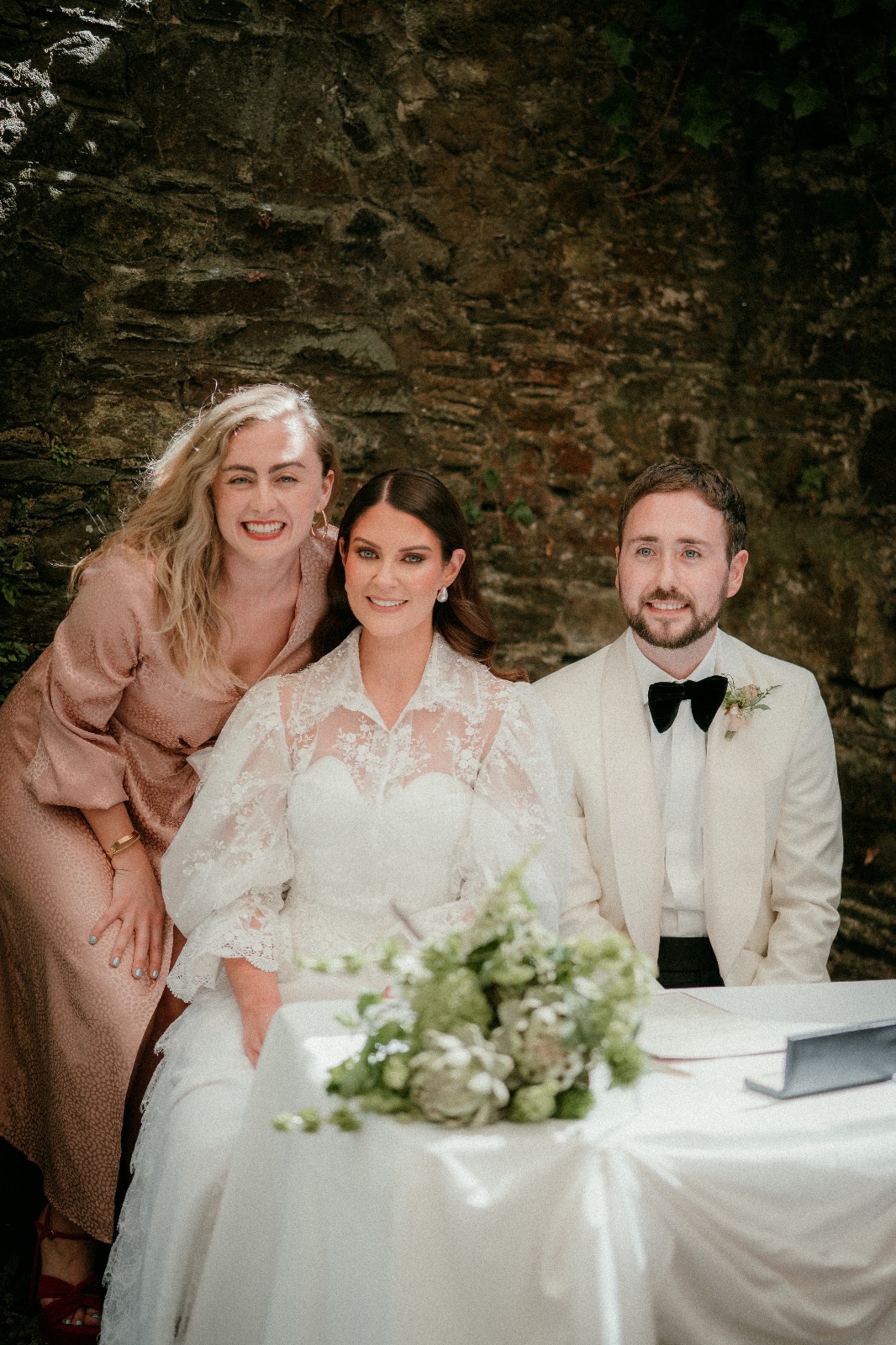Bride and groom sitting next to a guest in front of a brick wall, with a floral centerpiece on the table.