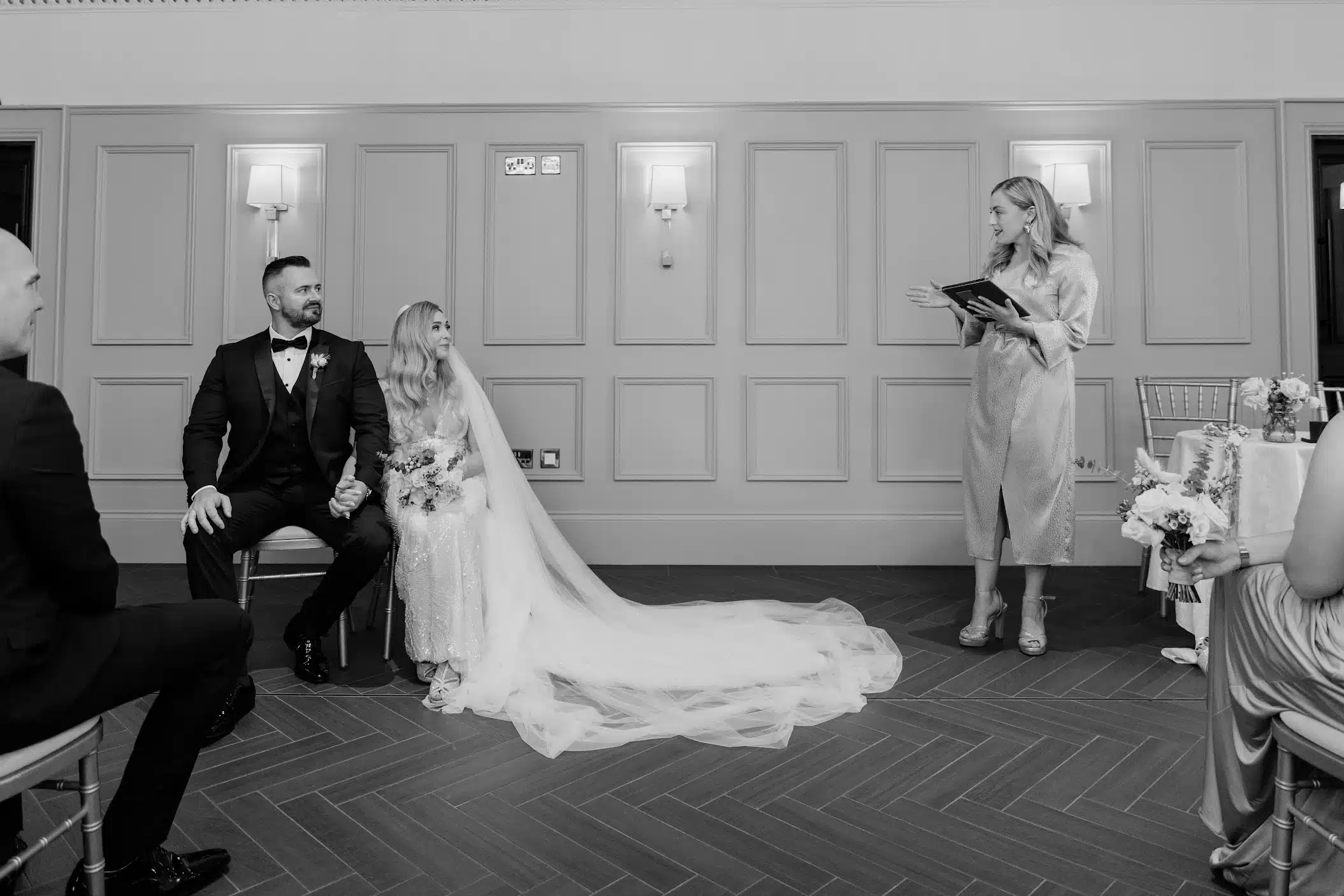 Bride and groom sitting during a wedding ceremony, with a woman officiating, in a formal setting.