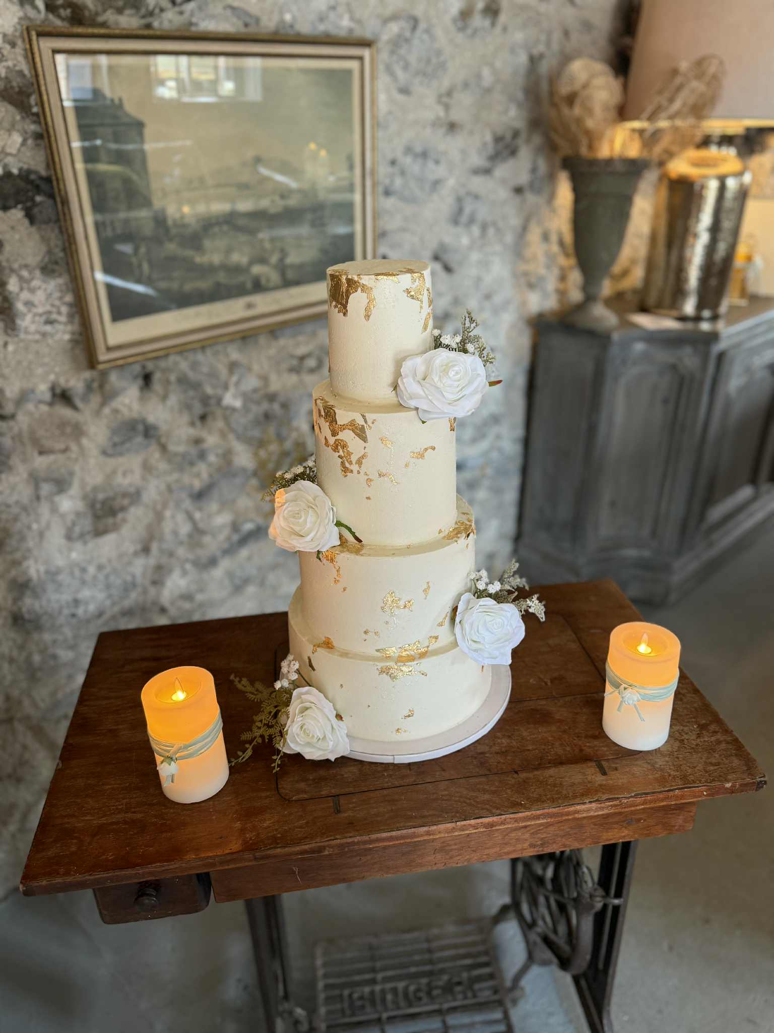 Four-tier cake with white roses and gold accents on a wooden table, surrounded by three candles, in a rustic setting.