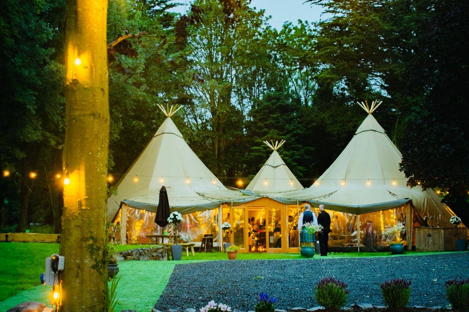 Illuminated tipi tents in a forest setting during an evening event.