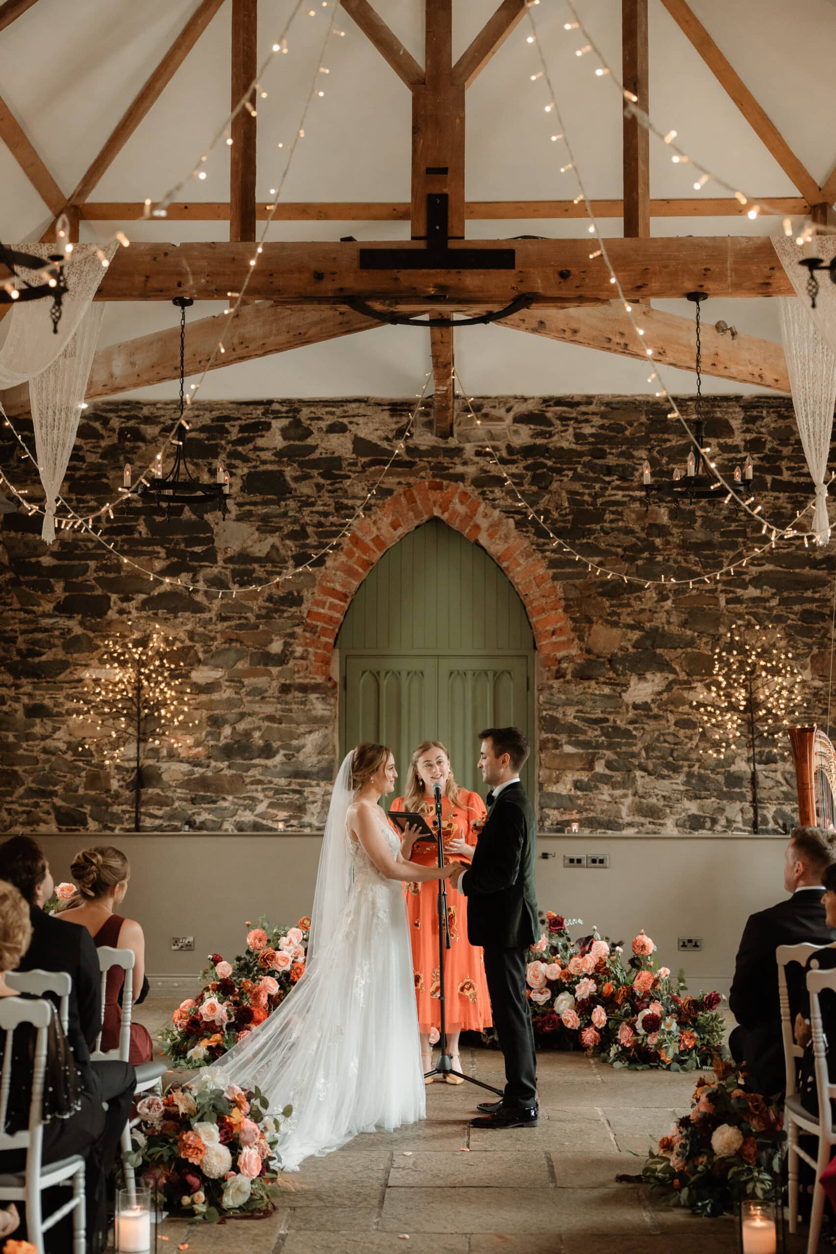 Bride and groom stand at altar in a rustic venue with stone walls, floral arrangements, and string lights.