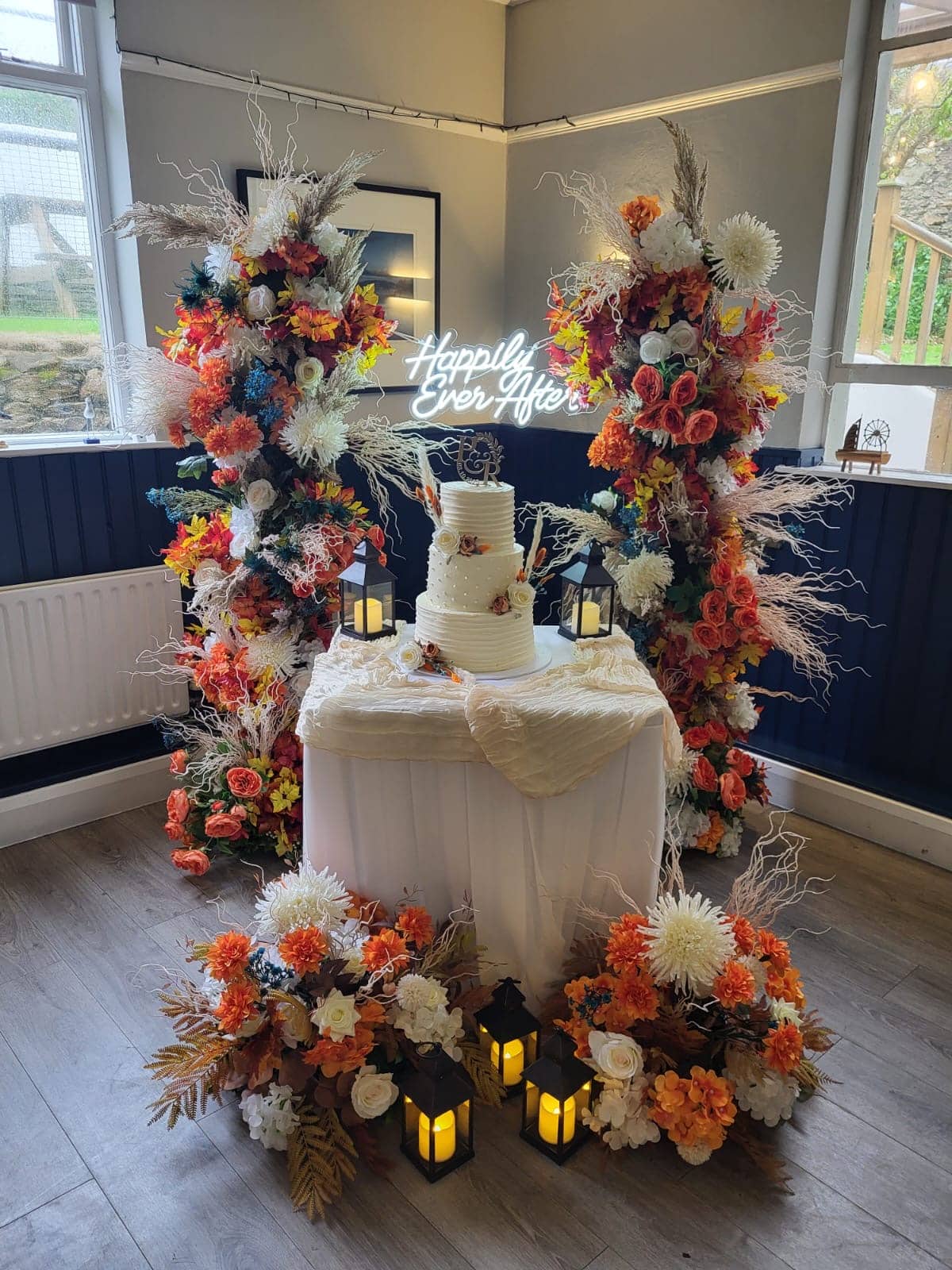 Wedding cake display with floral arrangements and a "Happily Ever After" sign, surrounded by lanterns and autumn-themed decor.