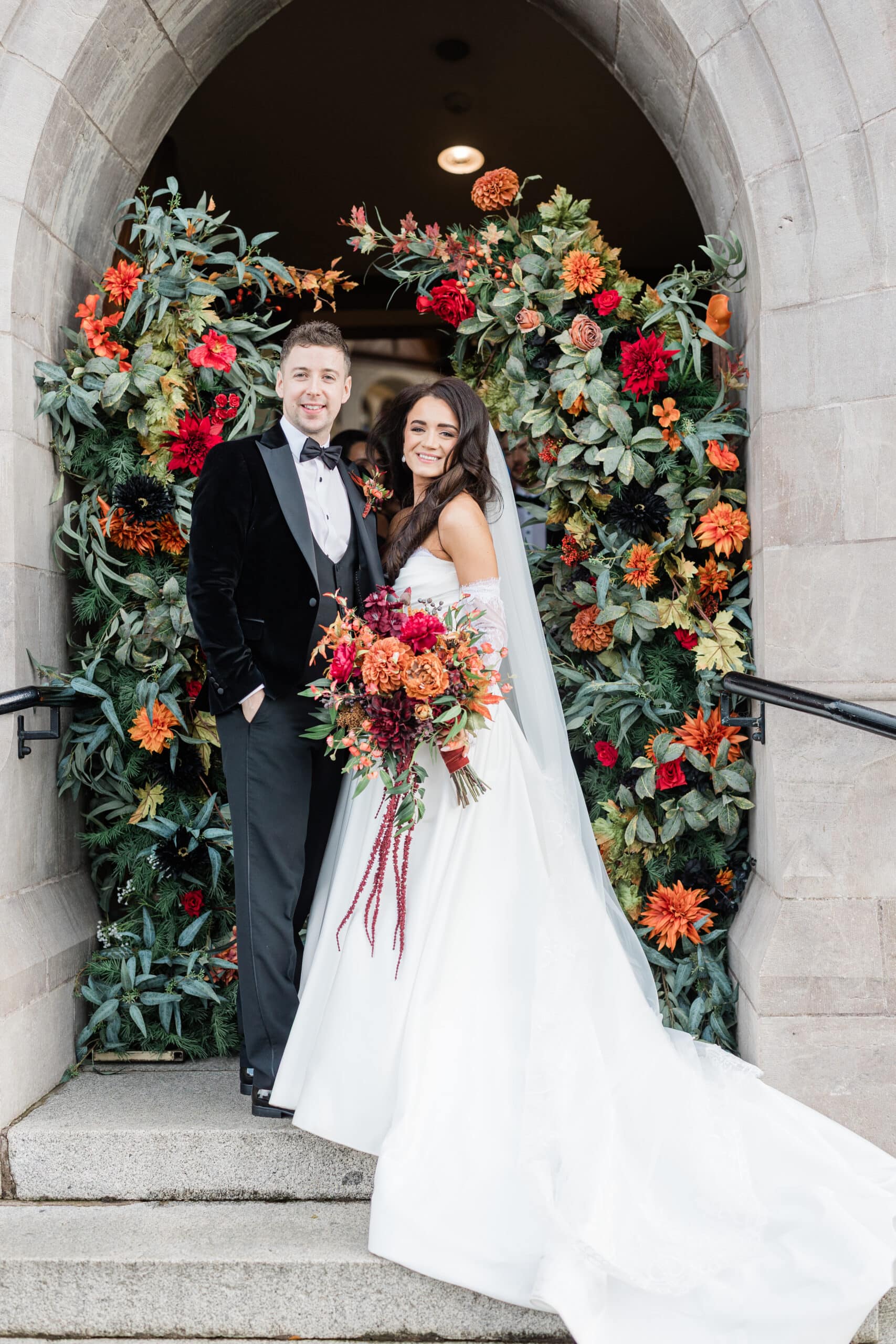 Bride and groom standing under floral arch at wedding, bride holding bouquet.