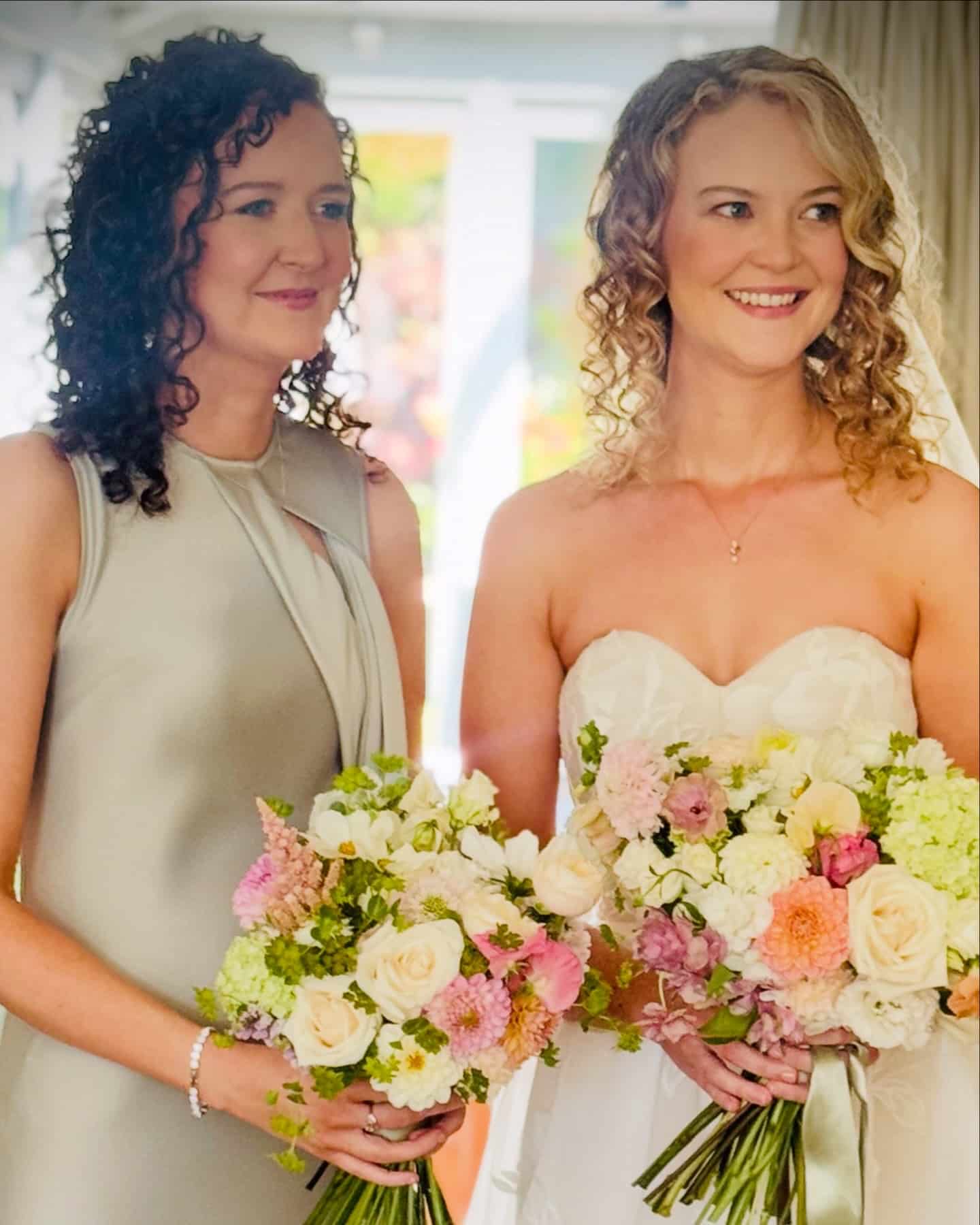 Two women smiling at a wedding, holding bouquets of colorful flowers.
