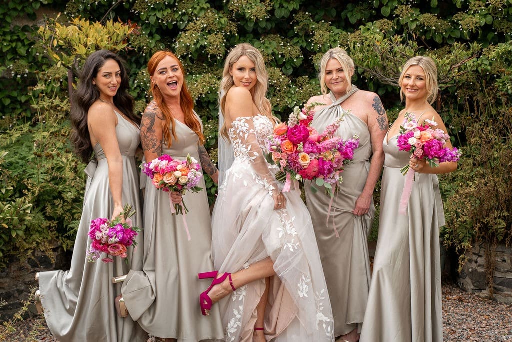 Bride and bridesmaids in silver dresses holding vibrant floral bouquets outdoors.