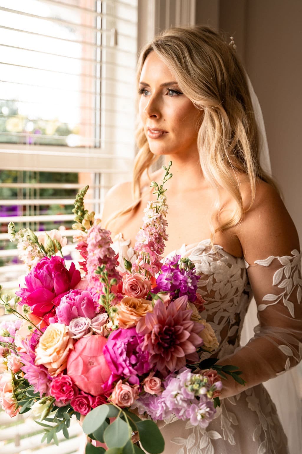 Bride with a vibrant bouquet of pink and peach flowers standing by a window.