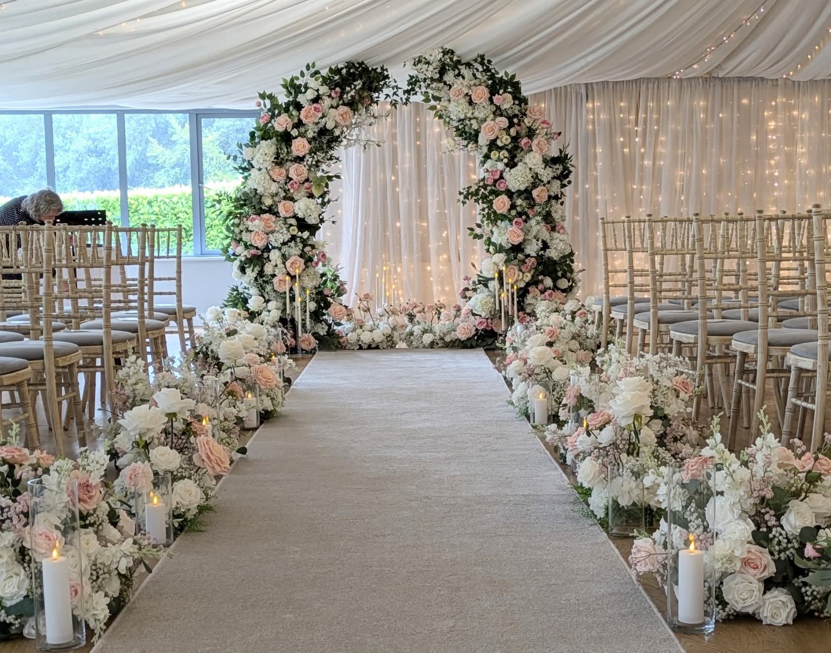 Elegant wedding venue with floral arch, aisle lined with flowers and candles, and rows of wooden chairs under a white tent.