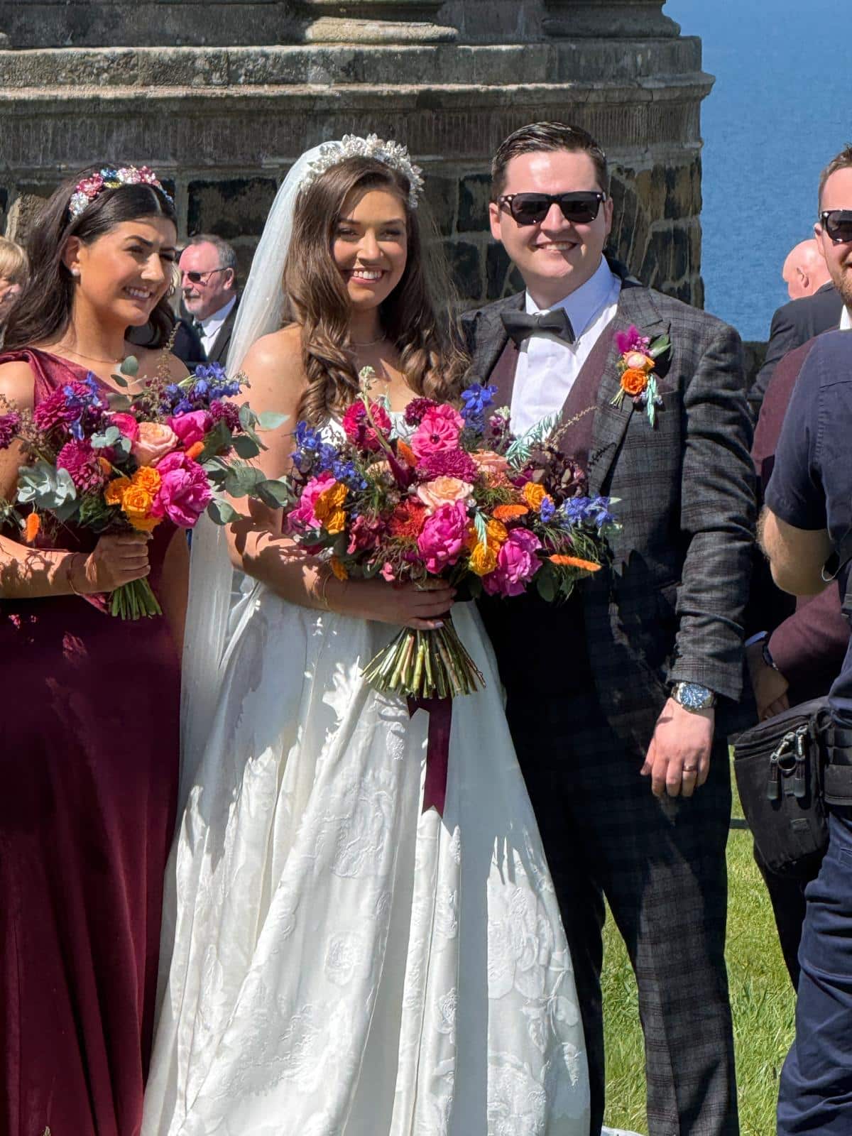 Bride and groom with bridesmaid holding bright floral bouquets at an outdoor wedding ceremony.