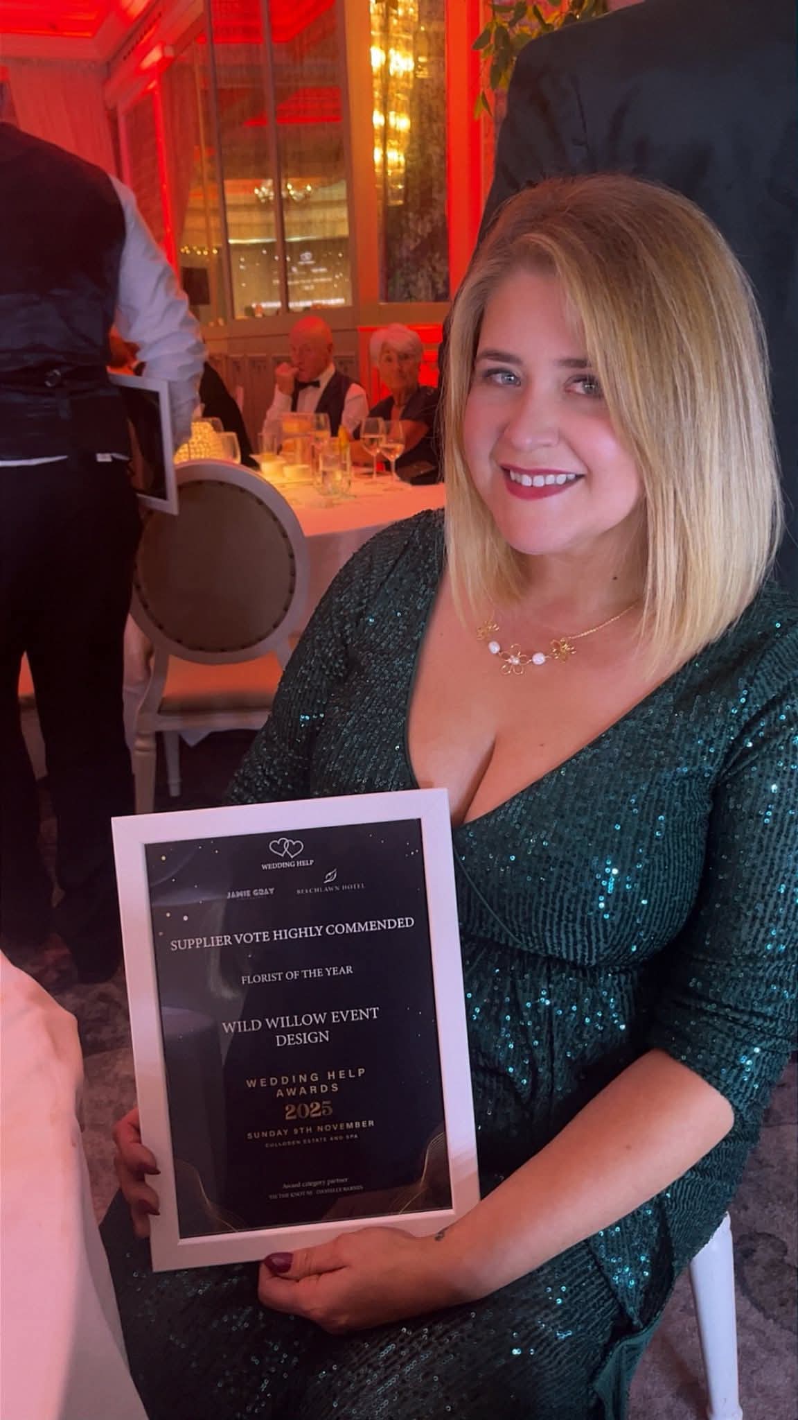 Woman holding an award at an event, seated at a table in a formal setting with red lighting.