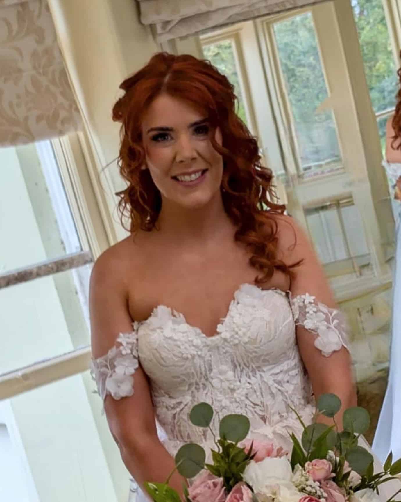 Bride with red hair in lace wedding dress holding bouquet in bright room.