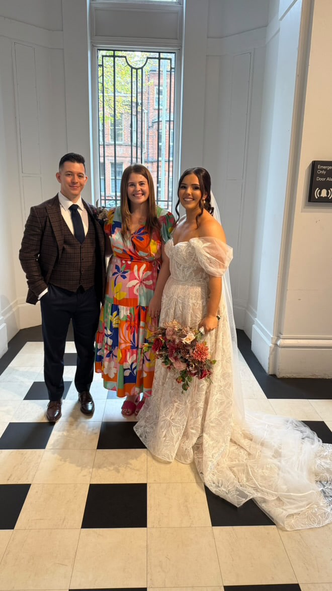 Bride in lace gown and veil with groom in suit and friend in colorful dress, posing indoors with a floral bouquet.