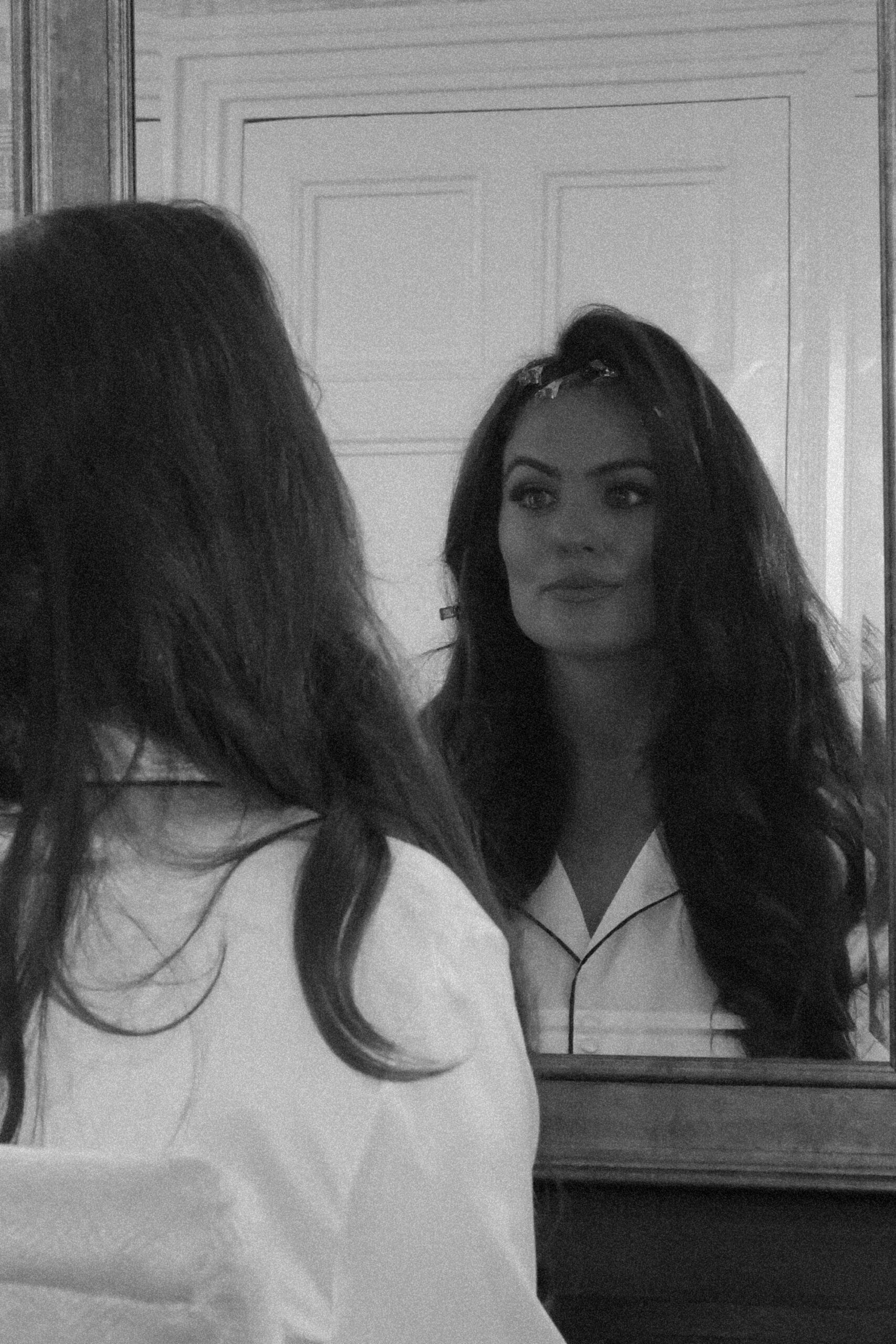 Black and white image of a woman looking at her reflection in the mirror, wearing a white shirt and hair clips.