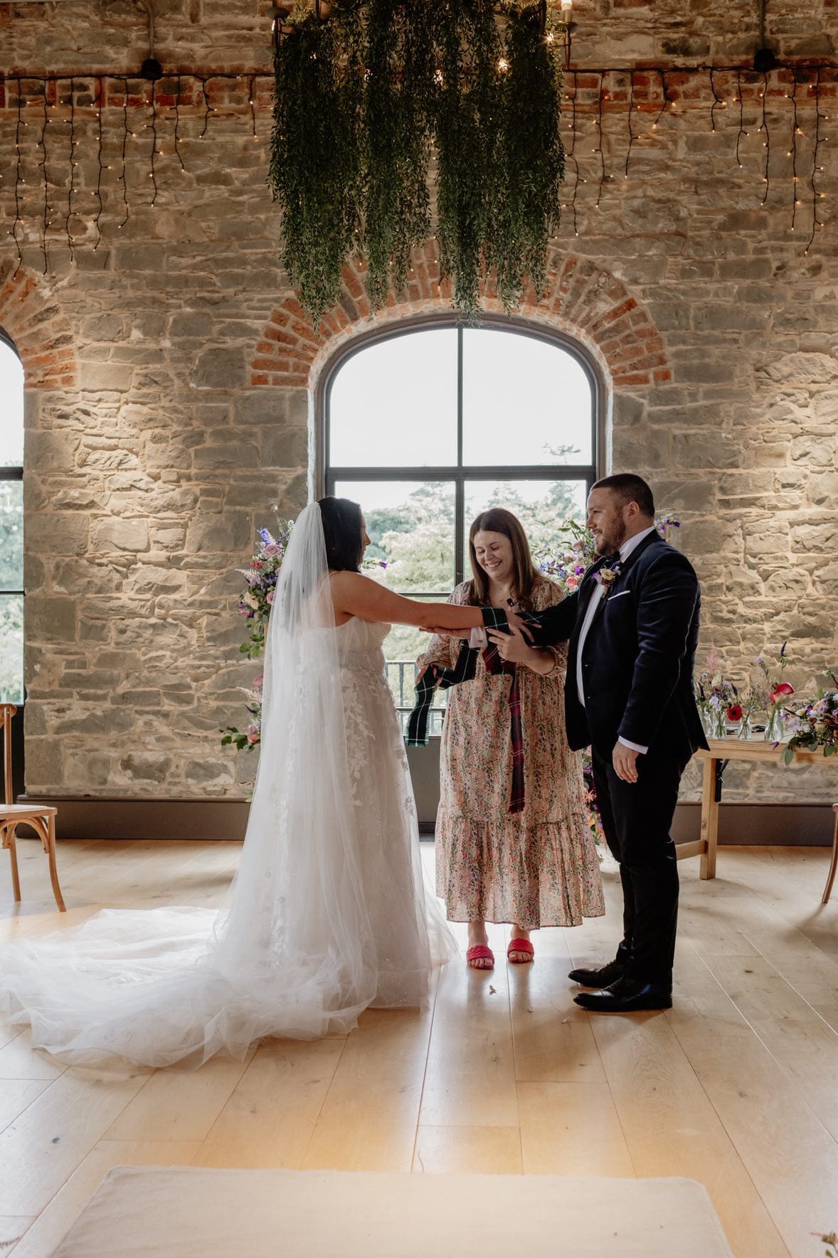 Bride and groom participate in a wedding ceremony with officiant in a rustic venue with brick walls and hanging greenery.