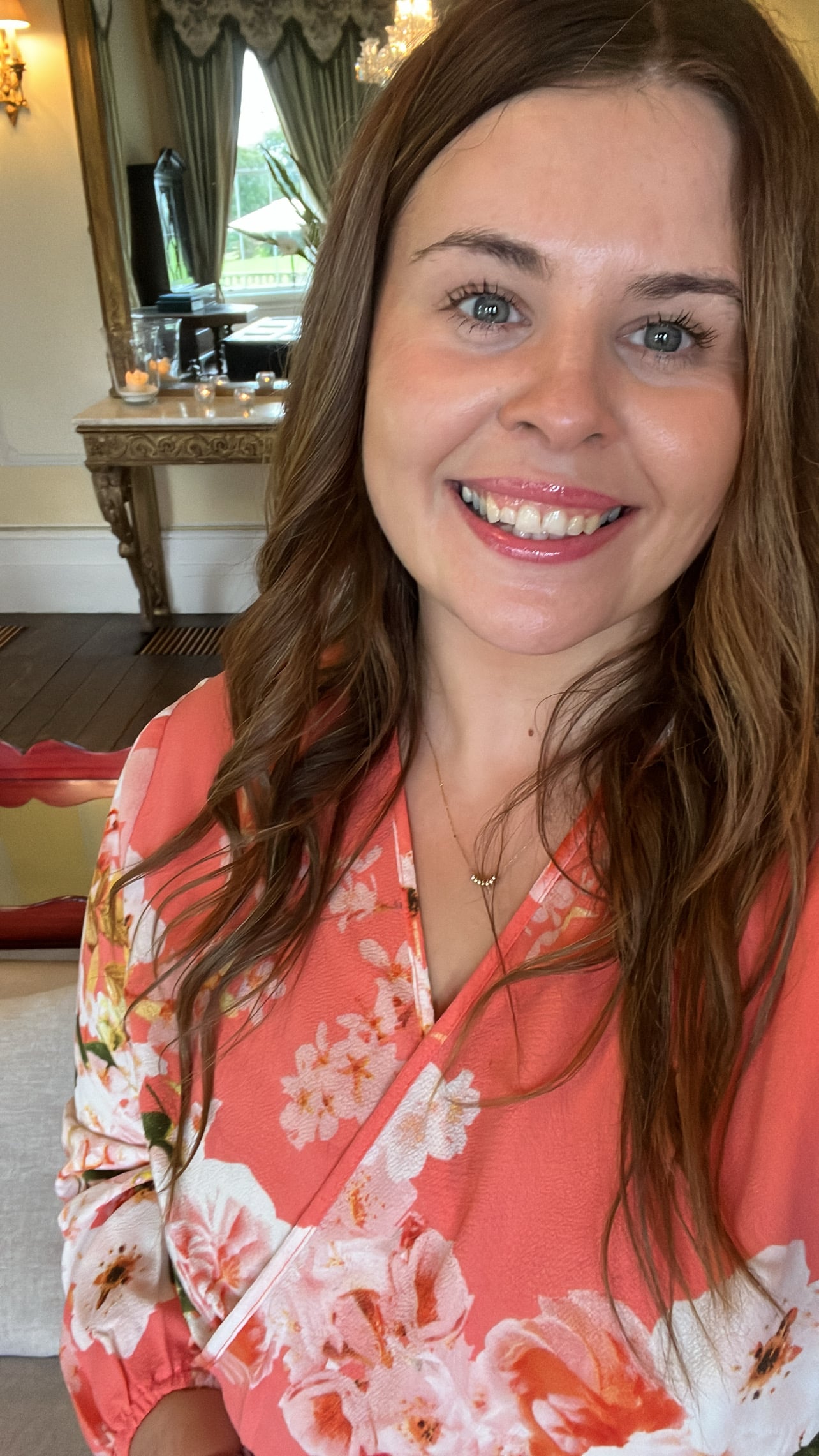 Smiling person in a floral pink dress, standing in an elegantly decorated room with chandeliers.
