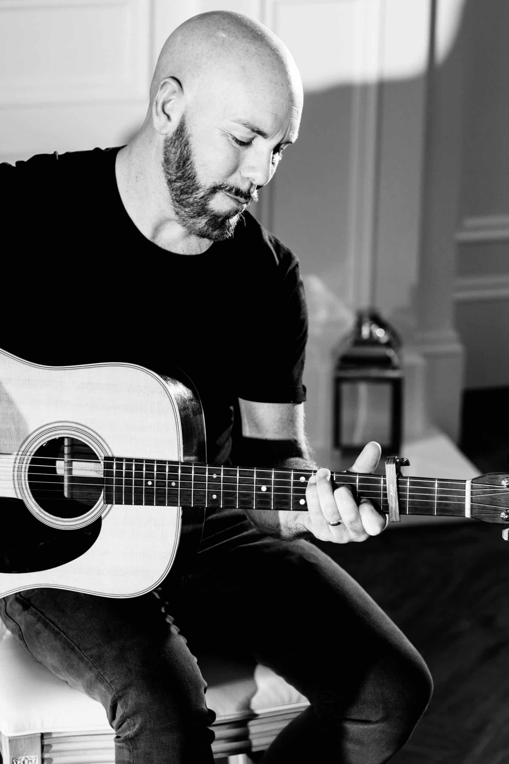 Bald man with a beard playing acoustic guitar indoors, in black and white.