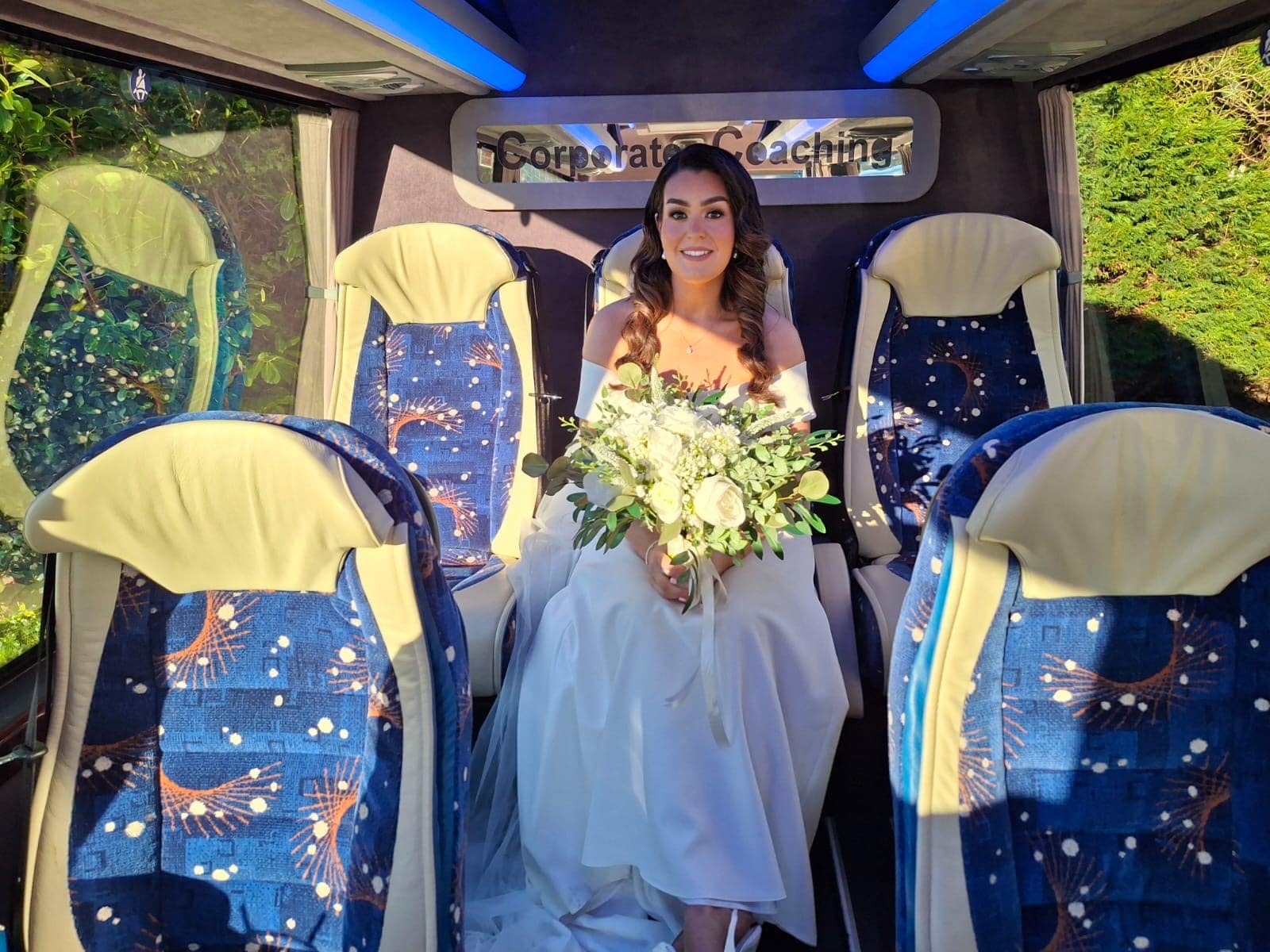 Bride holding bouquet seated in a decorative blue and beige bus interior with sunlit greenery outside.