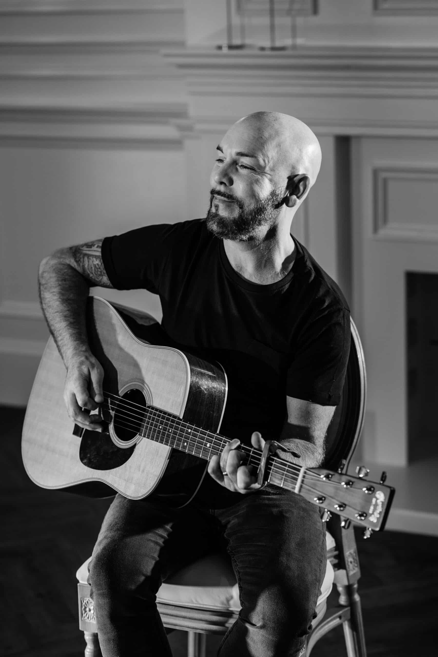 Bald man with a beard playing an acoustic guitar, seated indoors, black and white.
