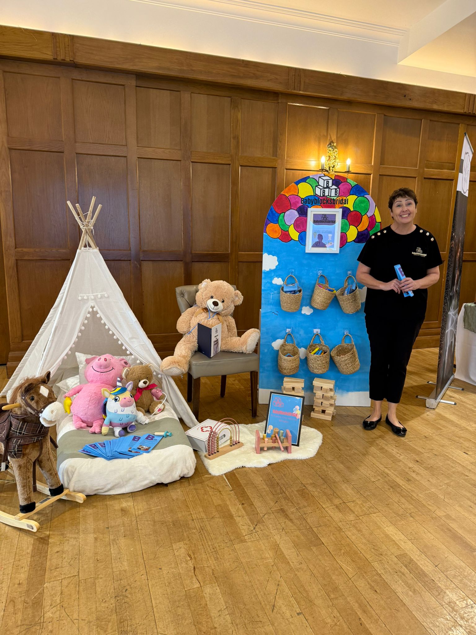 Toys and teepee display with a woman standing beside colorful backdrop in a wooden-paneled room.