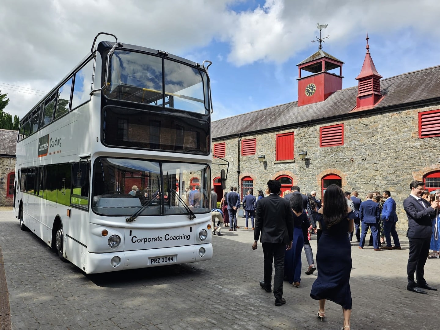 Double-decker bus parked outside a stone building with red doors, surrounded by people in formal attire.