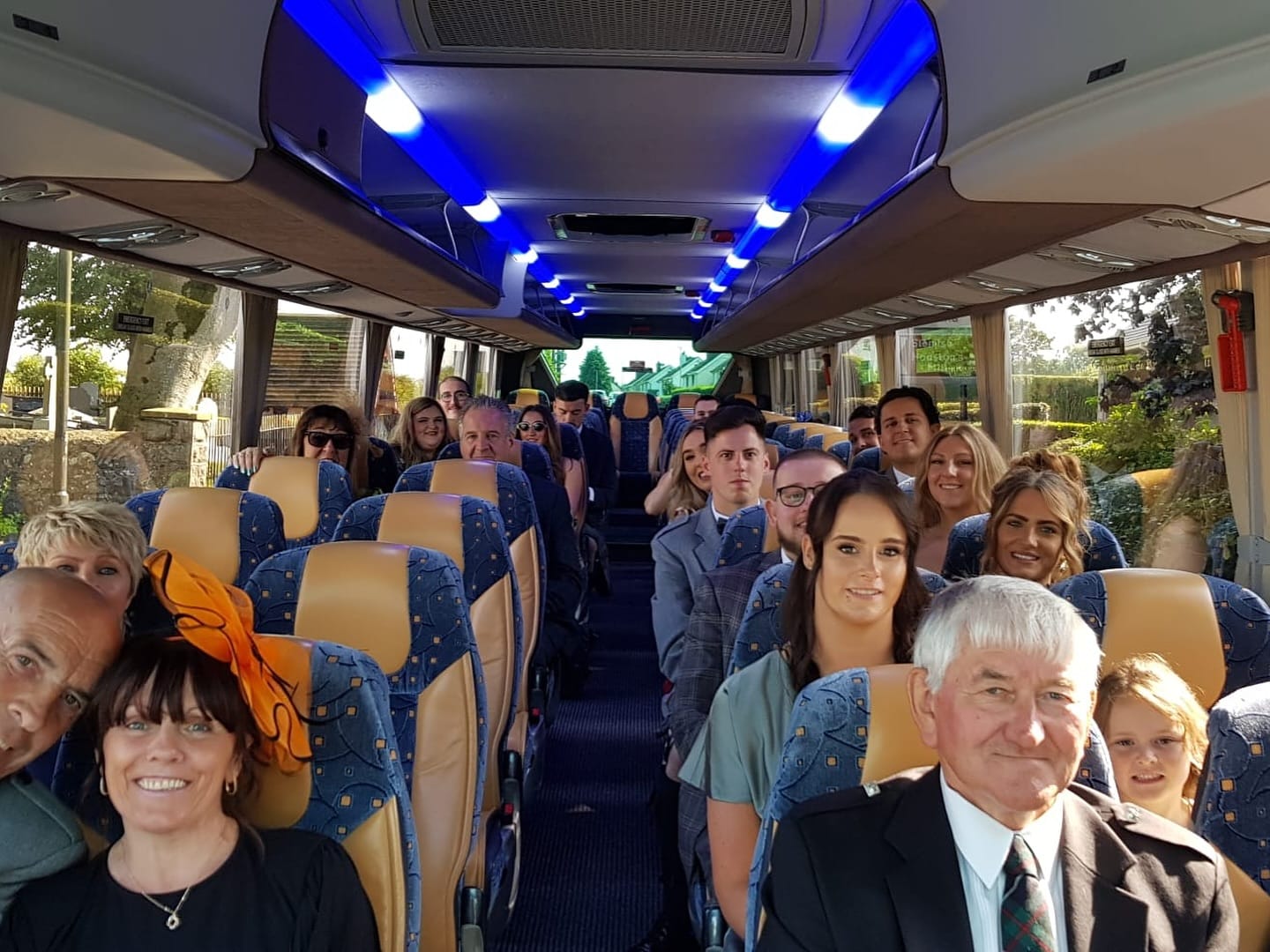 Group of people seated inside a bus, smiling at the camera, with blue lighting on the ceiling.