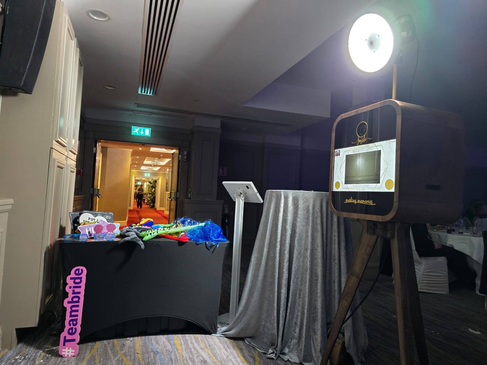 Photo booth setup with props on a table and a retro camera stand. Lit ring light and #TeamBride sign visible.