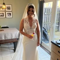 Bride in elegant wedding dress holding a drink, standing in a well-lit room with modern decor.