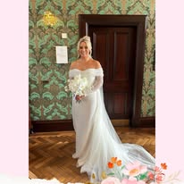 Bride in elegant white off-shoulder wedding gown holding bouquet indoors.