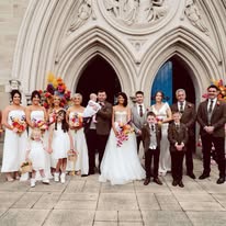 Wedding party posing in front of a church entrance, with bridesmaids, groomsmen, and children holding colorful bouquets.