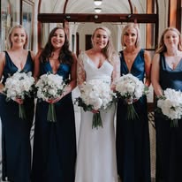 Bride and bridesmaids in navy dresses holding white bouquets in a hallway.