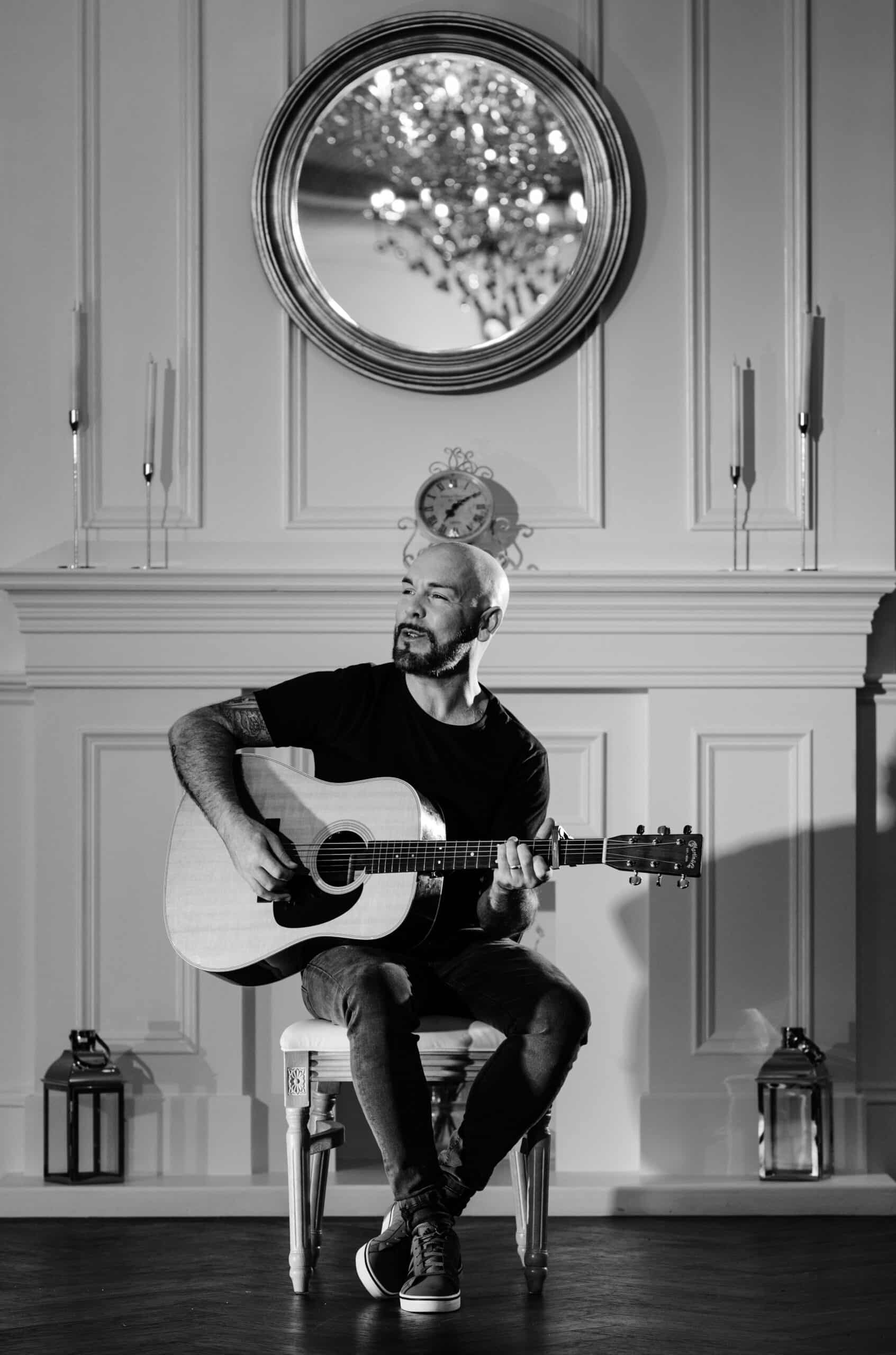 A man sitting and playing an acoustic guitar in a stylish room with a decorative mirror and candles.
