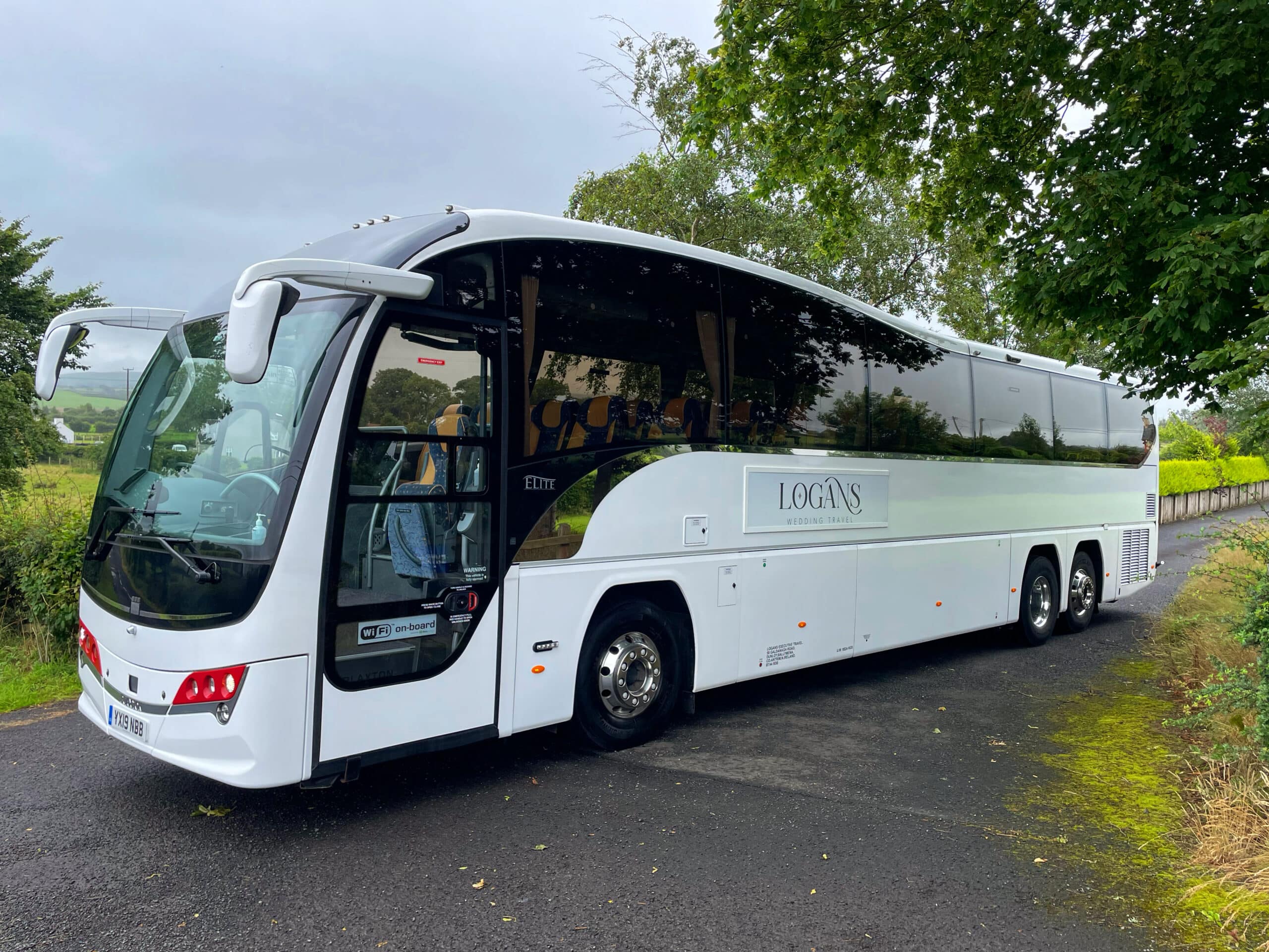 White tour bus parked on a road surrounded by greenery, labeled "Logan's" for wedding travel.