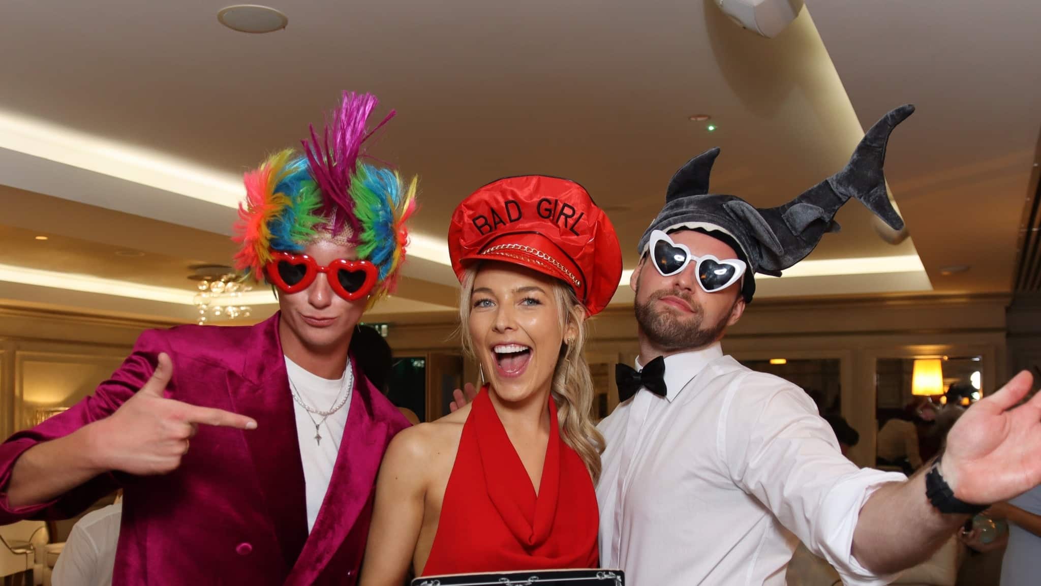Three people in colorful costumes and hats posing and smiling indoors at a party.