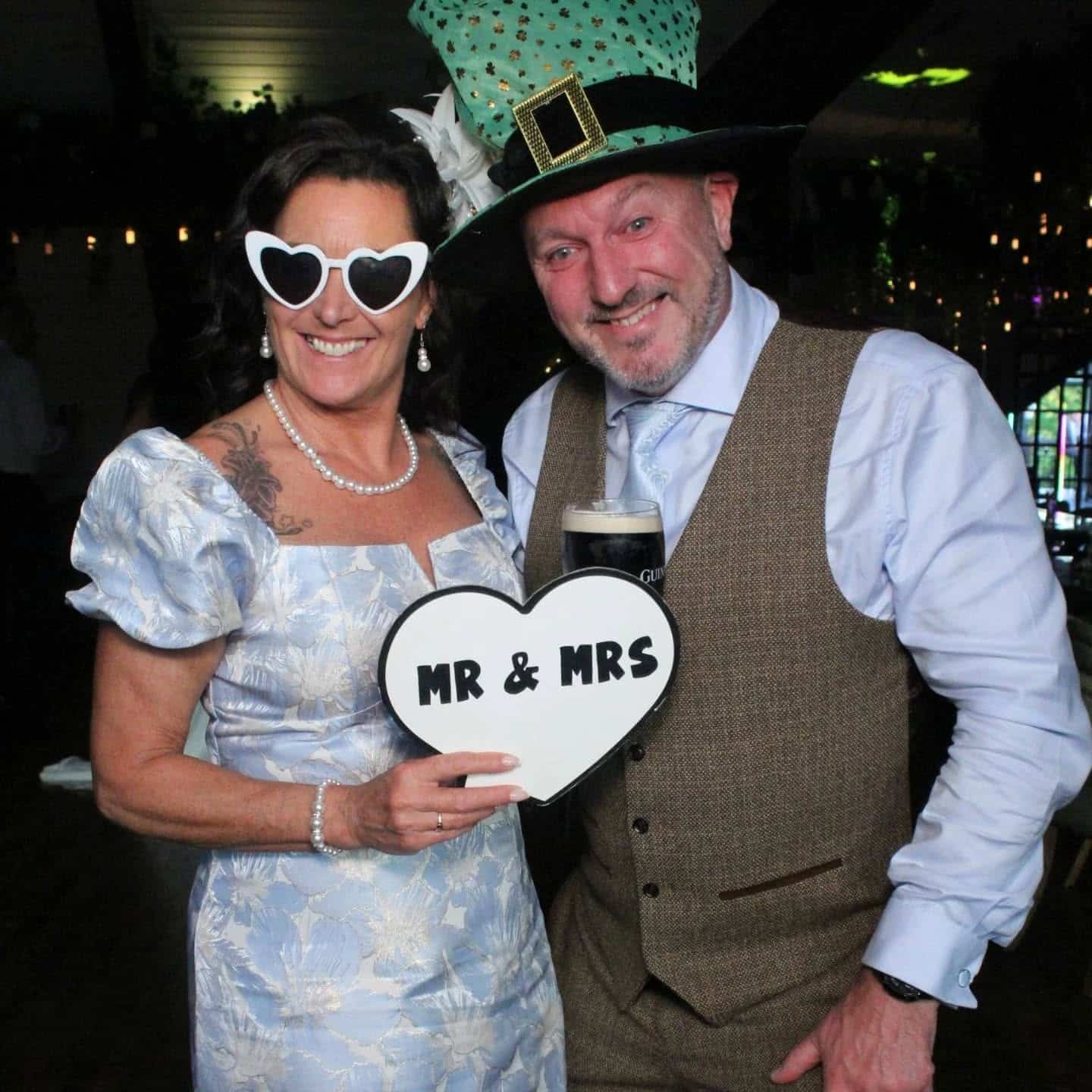 Couple dressed in festive attire, holding a "Mr & Mrs" heart sign, smiling at a celebration event.