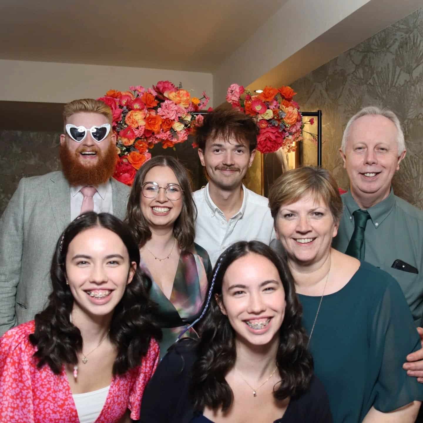 A group of people smiling at an indoor event with floral decorations in the background.