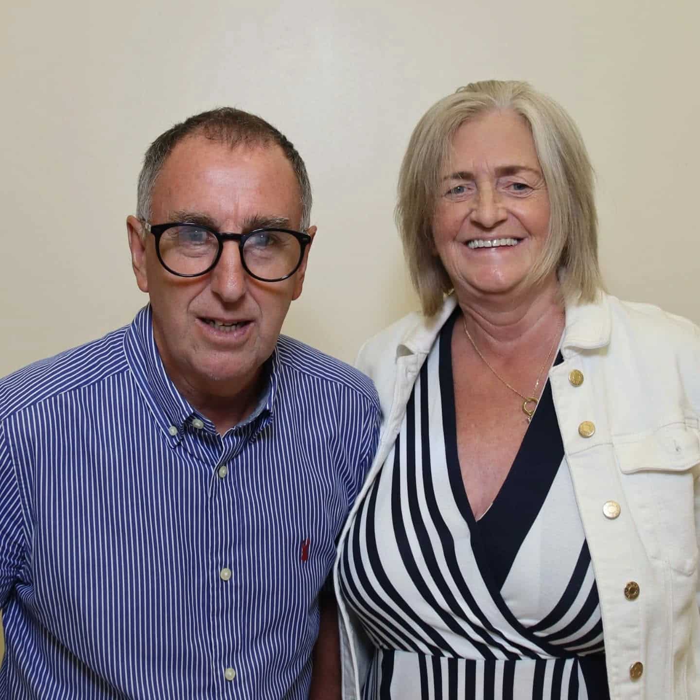 A smiling man in glasses and a striped shirt stands beside a woman in a striped dress and white jacket.