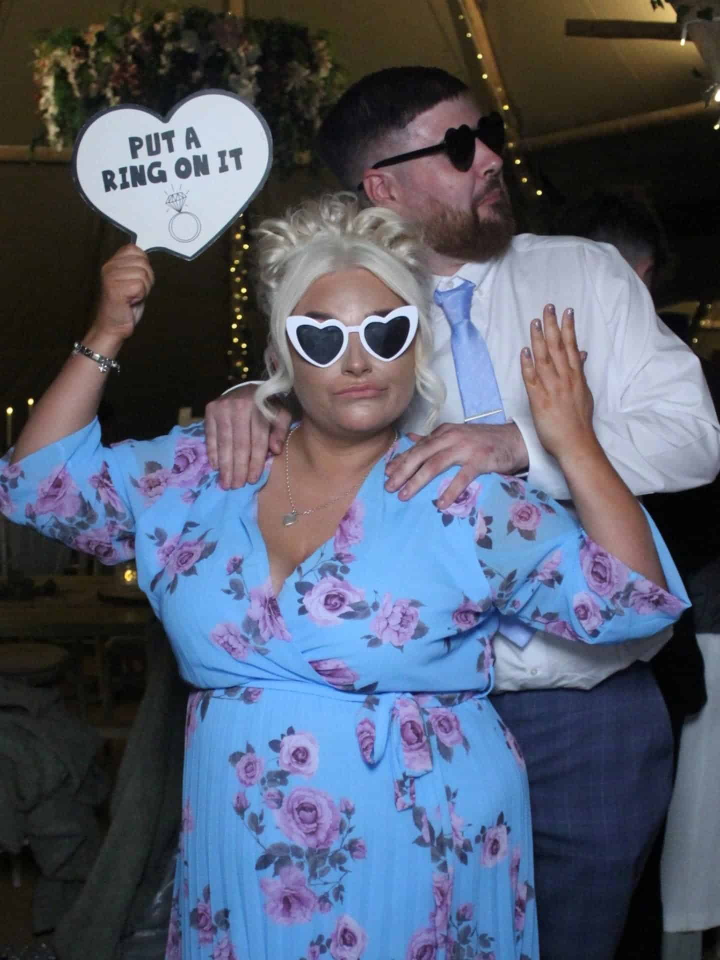 Couple wearing heart-shaped sunglasses, woman holding "Put a Ring on It" sign, posing playfully in floral-themed decor.